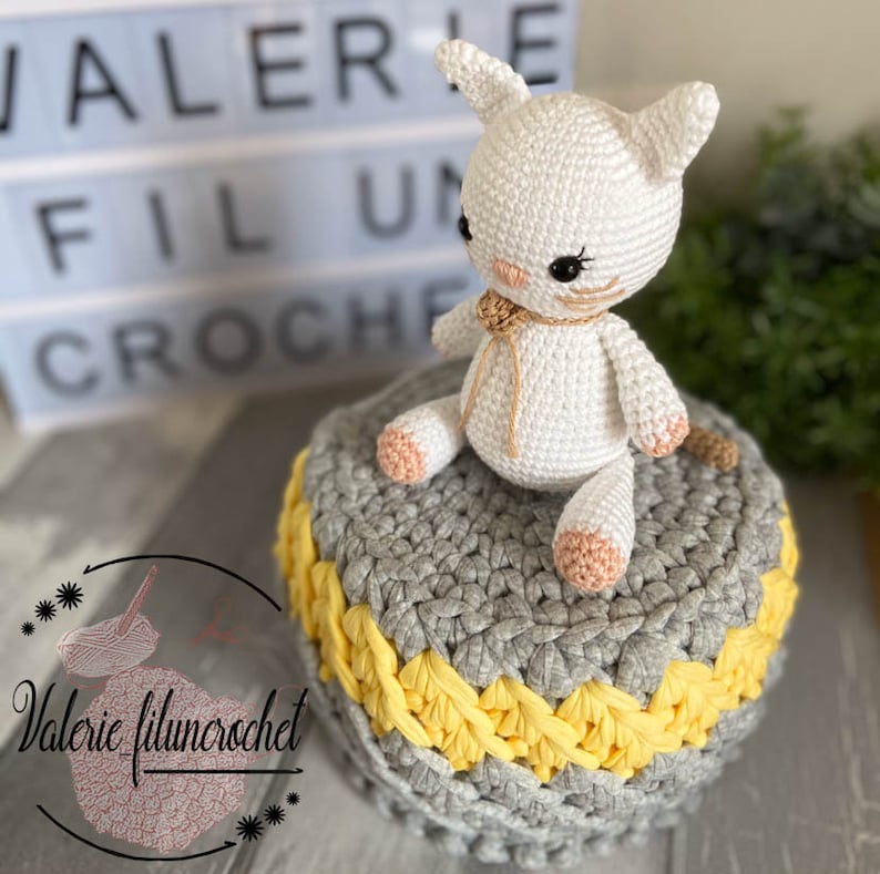 May include: A white crocheted cat with a yellow yarn bow sits on a grey and yellow crocheted stool. The stool has a grey base with a yellow crocheted trim.