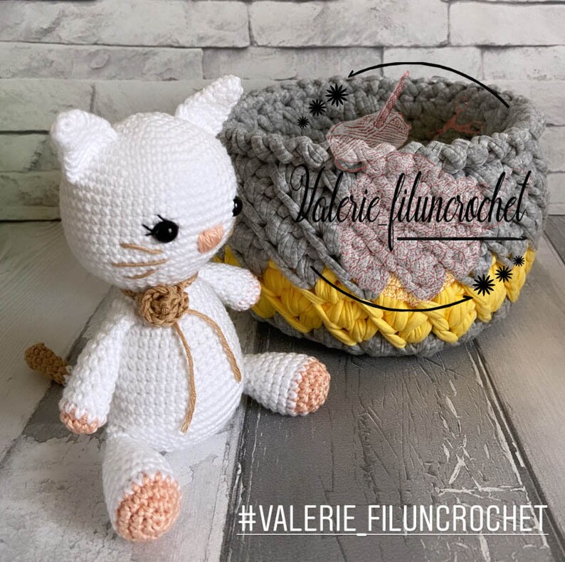 May include: A white crocheted cat toy with black eyes and a brown bow sits next to a grey, yellow, and pink crocheted basket. The basket has the text "Valerie filunccrochet" on it. The text "#VALERIE_FILUNCROCHET" is on the bottom of the image.