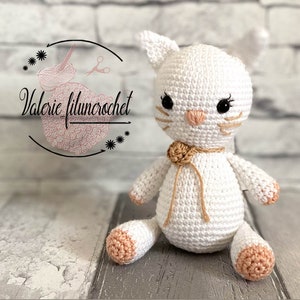 May include: A white crocheted cat with pink paws and a brown crocheted bow. The cat has black eyes and a pink nose.