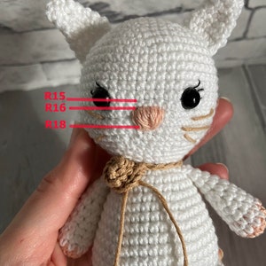 May include: White crocheted cat amigurumi with black eyes, pink nose, and brown yarn accents. The cat is labelled with the crochet row numbers R15, R16, and R18.