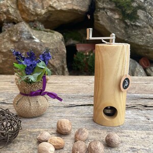 Könnte beinhalten: Ein hölzerner Muskatnuss-Mühle mit einer Handkurbel, ein kleiner Jutesack gefüllt mit lila Blumen und mehrere ganze Muskatnuss-Samen auf einer Holzoberfläche.