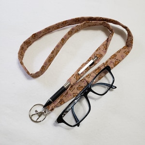 May include: A brown and tan fabric lanyard with a gingerbread man pattern. The lanyard has a silver clip and a black eyeglass chain attached to it. The eyeglass chain has a silver clip on one end and a black frame on the other.