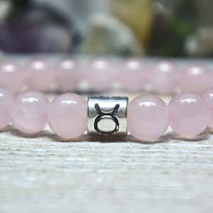 May include: A pink rose quartz bracelet with a silver charm featuring the Taurus zodiac symbol.