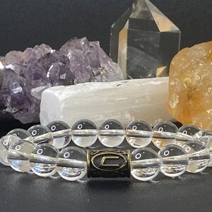 May include: A clear quartz crystal bead bracelet with a dark metal accent piece. The bracelet is displayed in front of a black background with amethyst, selenite, and other crystals.
