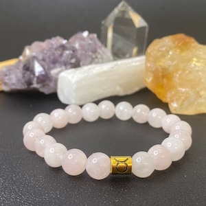 May include: A pink rose quartz beaded bracelet with a gold charm featuring the Taurus zodiac symbol.
