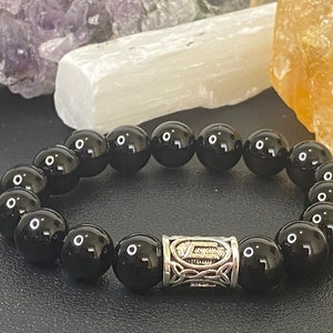 May include: A black beaded bracelet with a silver-colored spacer bead featuring an engraved design. The bracelet is made of smooth, round, polished black beads. The background includes crystals in shades of purple, white, and yellow.