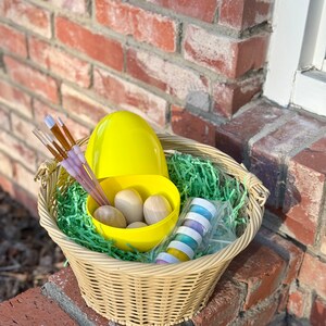 May include: A wicker basket filled with Easter craft supplies. The basket contains a large yellow egg, a yellow bowl with wooden eggs, paintbrushes, and small paint pots. Green paper grass fills the basket.