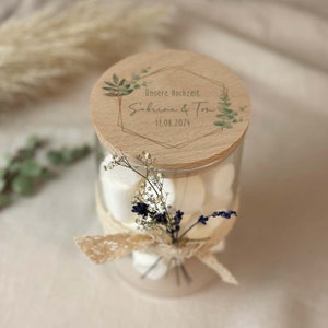 Könnte beinhalten: Ein Glasgefäß gefüllt mit weißen Marshmallows, verziert mit einem Holzdeckel mit floralem Design und dem Text "Unsere Hochzeit Sabrina & Tom 11.08.2024".