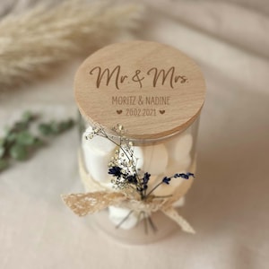 Könnte beinhalten: Ein Glasgefäß mit einem Holzdeckel, gefüllt mit weißen Marshmallows und mit getrockneten Blumen verziert. Der Deckel ist mit "Mr. & Mrs. Moritz & Nadine 2002.2021" und zwei kleinen Herzen graviert.