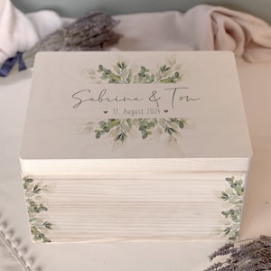 Puede incluir: Una caja de madera blanca con un diseño de hojas verdes y el texto "Sabrina & Tom" y "17. Agosto 2024".