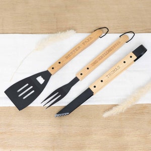 Può includere: Un set di tre utensili per grigliare con manici in legno. La spatola ha una lama nera con un apribottiglie. La forchetta ha denti neri. Le pinze hanno un bordo dentellato nero. I manici sono incisi con il testo "Bester Papa und Grillmeister Thomas".