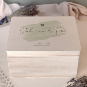 Puede incluir: Una caja de madera blanca con un diseño de acuarela verde y el texto "Sabrina & Tom" y "17. Agosto 2024".