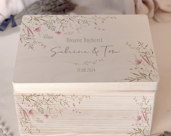 Op de afbeelding: Een witte houten doos met een bloemenontwerp en de tekst "Unsere Hochzeit Sabrina & Tom 11.08.2024" op de deksel.