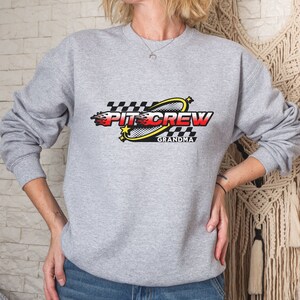 K&ouml;nnte beinhalten: Grauer Pullover mit einem schwarz-wei&szlig;en Karomuster, einem gelben Kreis mit einem roten und gelben Flammenmuster und dem Text "Pit Crew Grandma".