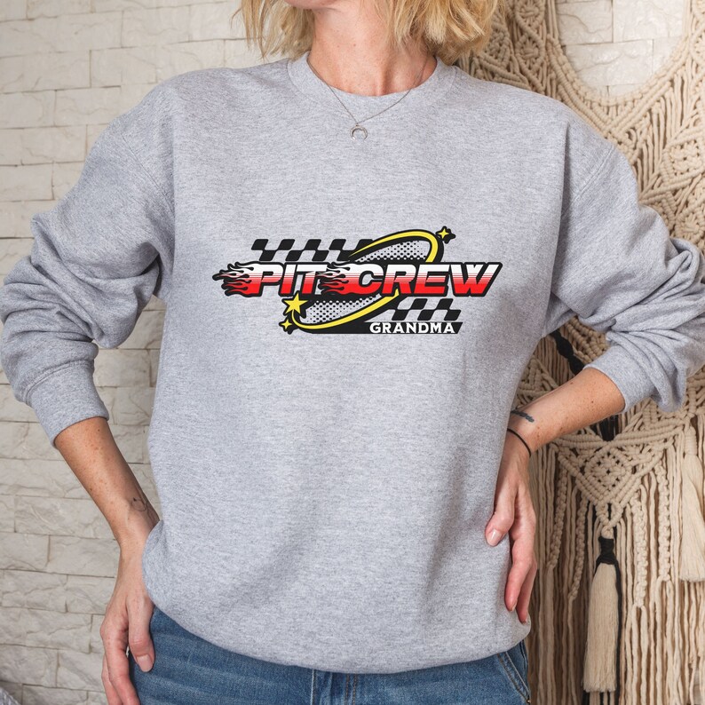 K&ouml;nnte beinhalten: Grauer Pullover mit einem schwarz-wei&szlig;en Karomuster, einem gelben Kreis mit einem roten und gelben Flammenmuster und dem Text "Pit Crew Grandma".