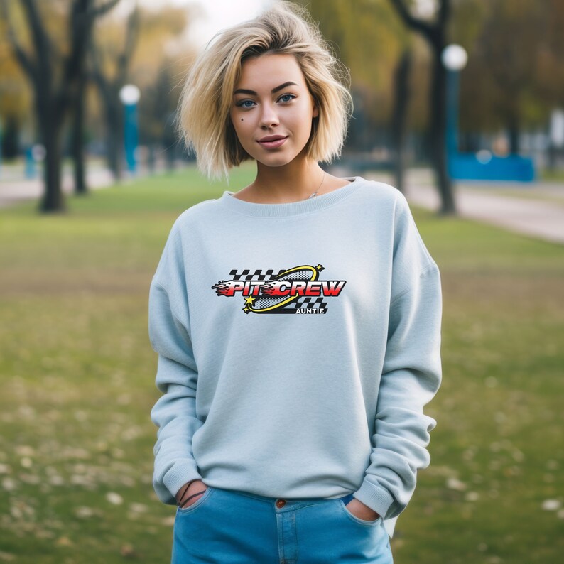 K&ouml;nnte beinhalten: Ein hellblaues Sweatshirt mit einem Grafikdesign aus einer karierten Flagge, einem gelben Stern, einer roten Flamme und dem Text "Pit Crew Auntie".