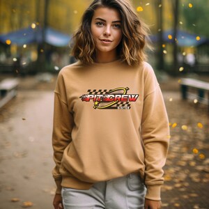 K&ouml;nnte beinhalten: Ein beiger Pullover mit einem Grafikdesign aus einem karierten Flaggenmuster, einer roten und gelben Flamme, einem gelben Kreis mit Sternen und dem Text "PIT CREW SISTER".