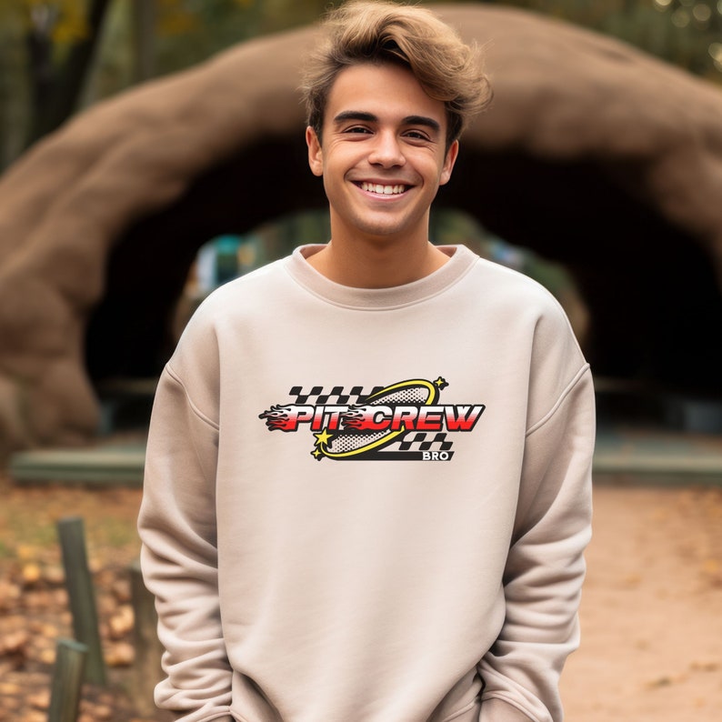 K&ouml;nnte beinhalten: Ein beiger Pullover mit einem grafischen Aufdruck eines karierten Flaggen, einem gelben Kreis und dem Text "PIT CREW BRO".