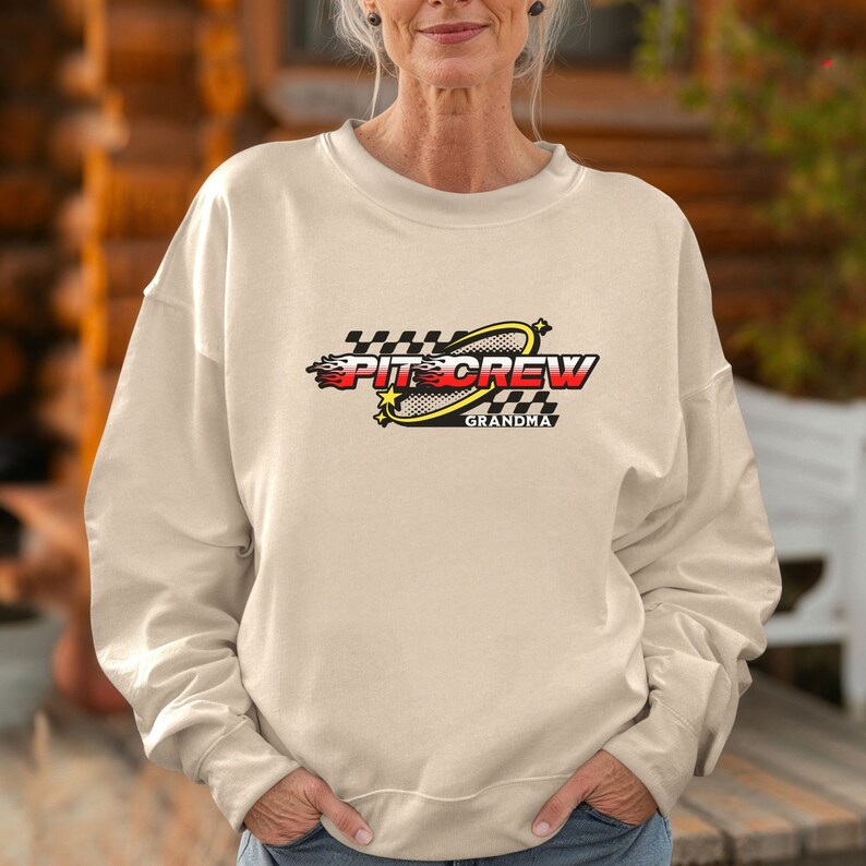 K&ouml;nnte beinhalten: Ein beiger Pullover mit einem grafischen Design eines Rennwagen-Boxencrew-Logos. Das Logo ist gelb, rot und schwarz mit dem Text "Pit Crew Grandma".