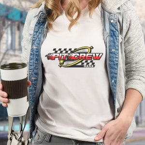 K&ouml;nnte beinhalten: Wei&szlig;es T-Shirt mit einem schwarzen, roten und gelben Grafikdesign, auf dem "Pit Crew Nana" steht. Die Grafik zeigt eine karierte Flagge, Flammen und einen Stern.