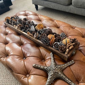 May include: A wooden tray filled with pine cones, dried orange slices, and cinnamon sticks. The tray is sitting on a brown leather ottoman with a silver starfish.