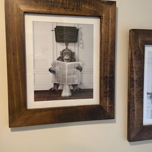 May include: A framed print showing a monkey on a toilet, engrossed in a newspaper. The print is set within a dark brown, rustic wooden frame. The image includes the text "MONKEY BUSINESS". A humorous and quirky piece of wall art.