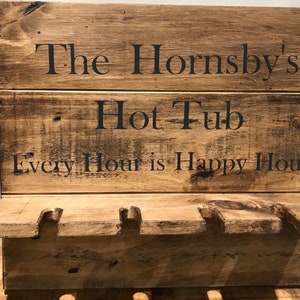 May include: A wooden sign with the text "The Hornsby's Hot Tub Every Hour is Happy Hour" in black lettering. The sign has a shelf with cutouts for glasses. The wood has a natural, aged appearance. A decorative item for a hot tub area.