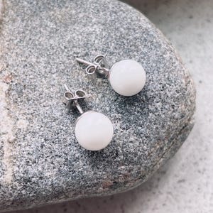 May include: A pair of white pearl stud earrings with silver posts. The earrings are displayed on a gray rock.