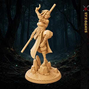 May include: A tan-colored miniature figurine of a warrior standing on a rock formation base with a circular, rippled pattern. The figure holds a long staff, wears wrapped clothing, and is posed in a dynamic stance with one leg raised.