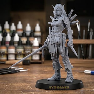 May include: A grey figurine of a female warrior, holding a sword and a dagger. The figure is detailed with armour, pouches, and boots, and is mounted on a black base with the text "SYZ GAMES". The background includes paint bottles and brushes.