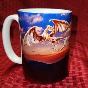 Könnte beinhalten: Weiße Keramiktasse mit einem bunten Bild eines Drachen, der in einem Sonnenuntergangshimmel fliegt. Der Drache ist braun und golden vor einem blauen und violetten Hintergrund.