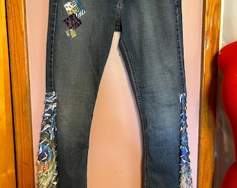 May include: Dark wash denim jeans with decorative patches and fabric accents. The jeans feature a patchwork design on the left thigh and flowing fabric trim along the outer seams, with a mix of blue, green, and purple hues.