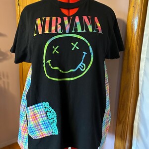 May include: Black t-shirt with a tie-dye "NIRVANA" graphic and a smiley face with X's for eyes. The shirt has a cut-out neckline and is layered with a plaid fabric.