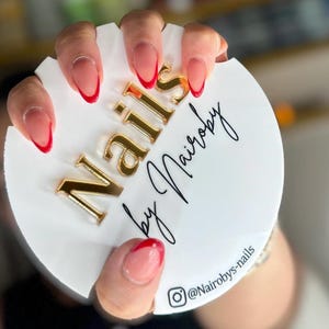 May include: Close-up of a hand with almond-shaped nails painted with a sheer pink polish and a thin red line at the tip. The hand is holding a white round sign with gold lettering that reads "Nails by Nairobi" and a social media handle "@Nairobiys-nails".