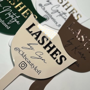 May include: Three different shaped, white, brown, and green signs with the word "LASHES" in black lettering. Each sign has a different name written in cursive below the word "LASHES". The signs have a social media handle and an Instagram icon.