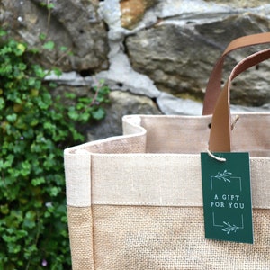 An up-close of our Hessian Market Shopper bag in front of a rustic wall. A green 'Boutique Favours' gift tag is attached.