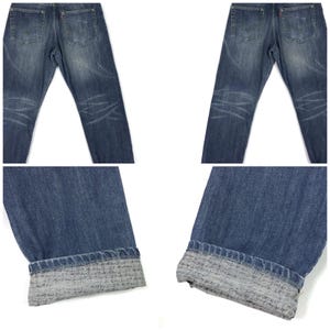 May include: A pair of blue denim jeans with a cuffed hem. The jeans have a faded and distressed look, with visible wear and tear. The cuffs are turned up to reveal a gray and white patterned fabric.