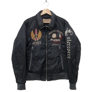 Puede incluir: Chaqueta bomber acolchada negra con ribete gris y parches bordados. Presenta el texto "Postes France" y "Avirex Europa". Incluye cierre de cremallera y dos bolsillos delanteros. La chaqueta est&aacute; colgada en una percha de madera.