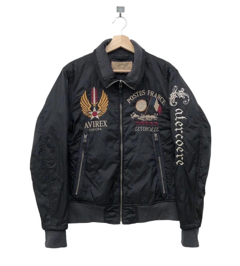 Puede incluir: Chaqueta bomber acolchada negra con ribete gris y parches bordados. Presenta el texto "Postes France" y "Avirex Europa". Incluye cierre de cremallera y dos bolsillos delanteros. La chaqueta est&aacute; colgada en una percha de madera.