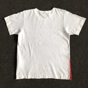 Puede incluir: Camiseta blanca con un dise&ntilde;o negro y rojo en las costuras laterales.