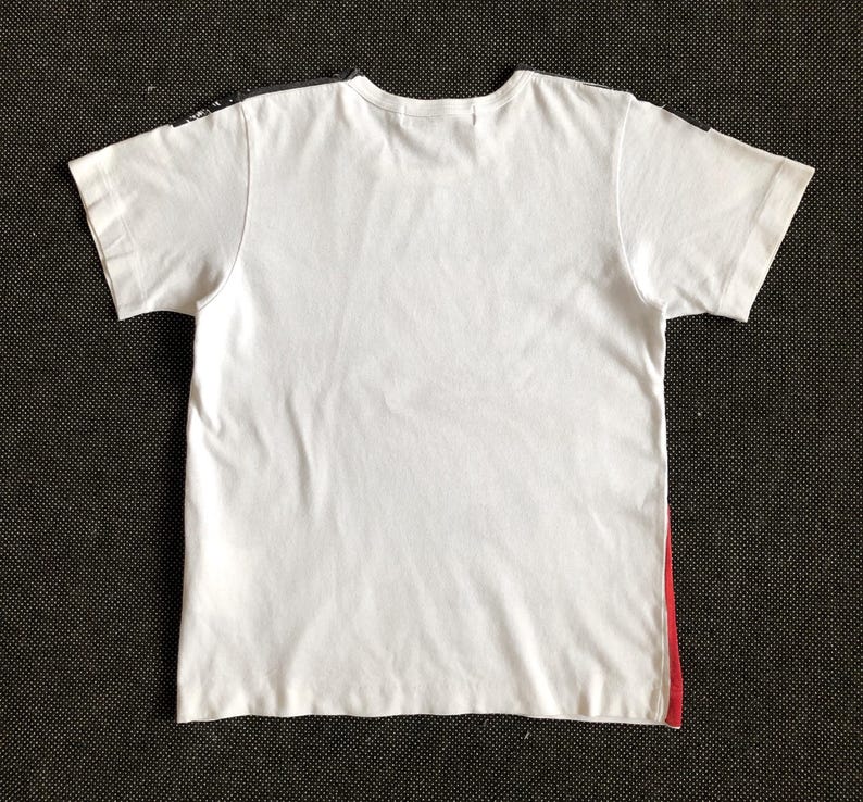 Puede incluir: Camiseta blanca con un dise&ntilde;o negro y rojo en las costuras laterales.