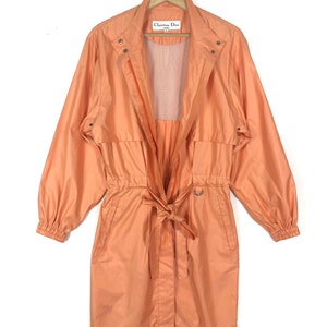 May include: A long-sleeved, peach-coloured jacket with a zip closure, a drawstring waist, and a label that reads "Christian Dior" on the left side of the collar.
