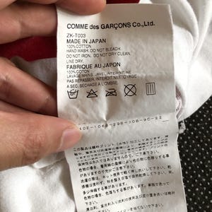 Puede incluir: Etiqueta de cuidado de la ropa blanca con texto negro que dice "COMME des GAR&Ccedil;ONS Co., Ltd." y "ZK-T003 MADE IN JAPAN 100% COTTON HAND WASH. DO NOT BLEACH. DO NOT IRON. DO NOT DRY CLEAN. LINE DRY. FABRIQUE AU JAPON 100% COTON LAVAGE MAINS. JAVEL INTERDIT. NE PAS REPASSER. INTERDIT NETTOYAGE A SEC. SECHAGE A L'OMBRE." La etiqueta tambi&eacute;n incluye cuatro s&iacute;mbolos: un cubo con agua, una plancha con una X tachada, un c&iacute;rculo con una X tachada y un cuadrado con tres l&iacute;neas verticales.