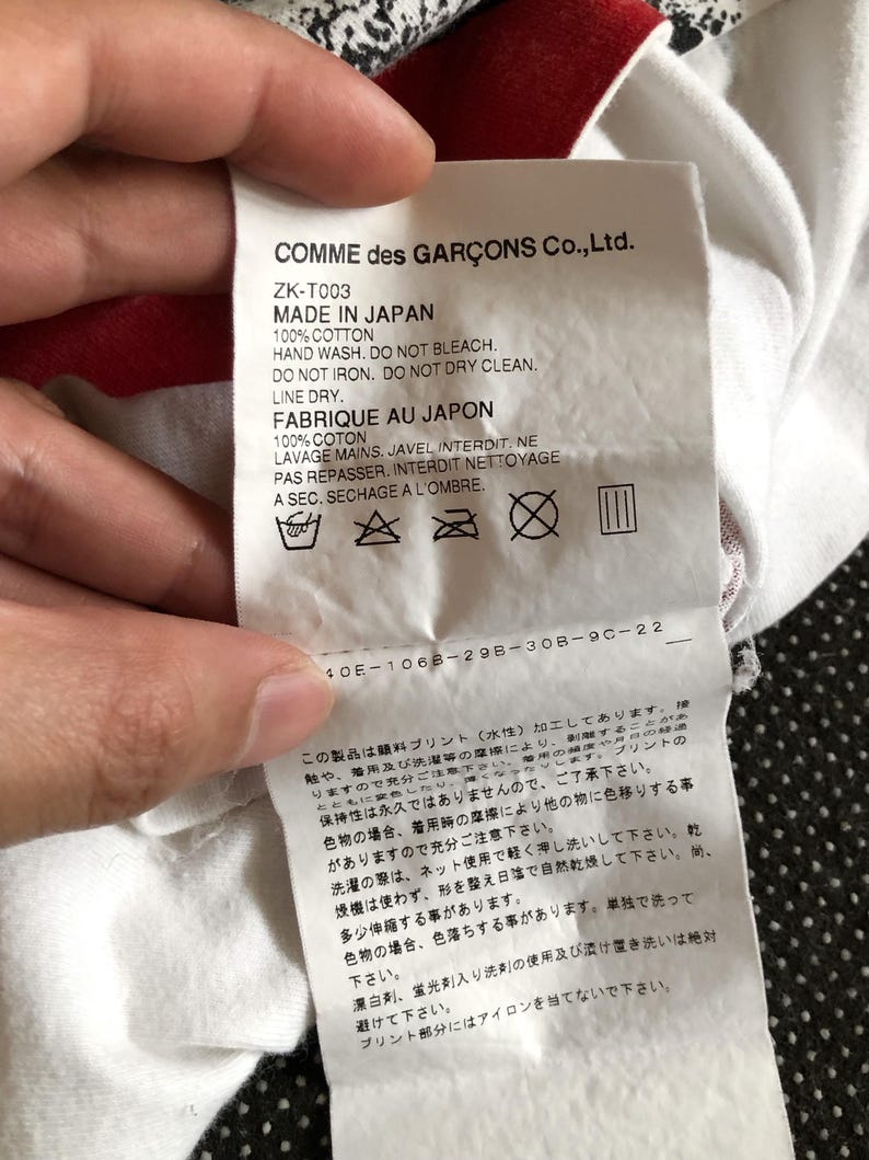 Puede incluir: Etiqueta de cuidado de la ropa blanca con texto negro que dice "COMME des GAR&Ccedil;ONS Co., Ltd." y "ZK-T003 MADE IN JAPAN 100% COTTON HAND WASH. DO NOT BLEACH. DO NOT IRON. DO NOT DRY CLEAN. LINE DRY. FABRIQUE AU JAPON 100% COTON LAVAGE MAINS. JAVEL INTERDIT. NE PAS REPASSER. INTERDIT NETTOYAGE A SEC. SECHAGE A L'OMBRE." La etiqueta tambi&eacute;n incluye cuatro s&iacute;mbolos: un cubo con agua, una plancha con una X tachada, un c&iacute;rculo con una X tachada y un cuadrado con tres l&iacute;neas verticales.