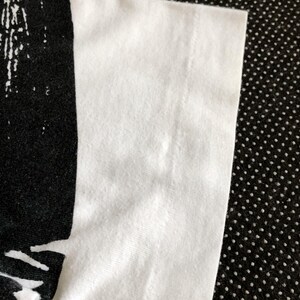 Puede incluir: Primer plano del tejido de algod&oacute;n blanco de una camiseta. El tejido es liso y tiene un ligero brillo. El borde del tejido est&aacute; doblado y cosido.