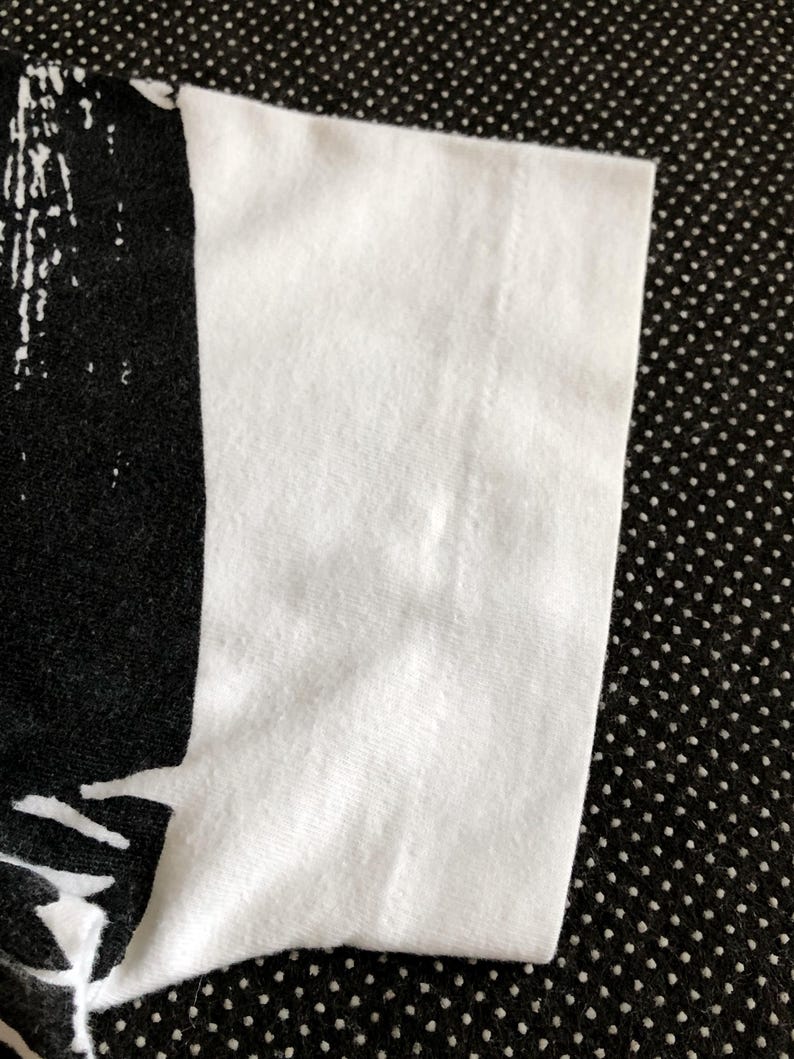 Puede incluir: Primer plano del tejido de algod&oacute;n blanco de una camiseta. El tejido es liso y tiene un ligero brillo. El borde del tejido est&aacute; doblado y cosido.