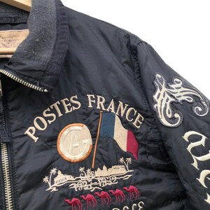 Puede incluir: Chaqueta acolchada azul marino con bordado "POSTES FRANCE", bandera francesa y figuras de camellos. Cuello gris y cierre de cremallera. Parches decorativos con letras blancas en la manga derecha.