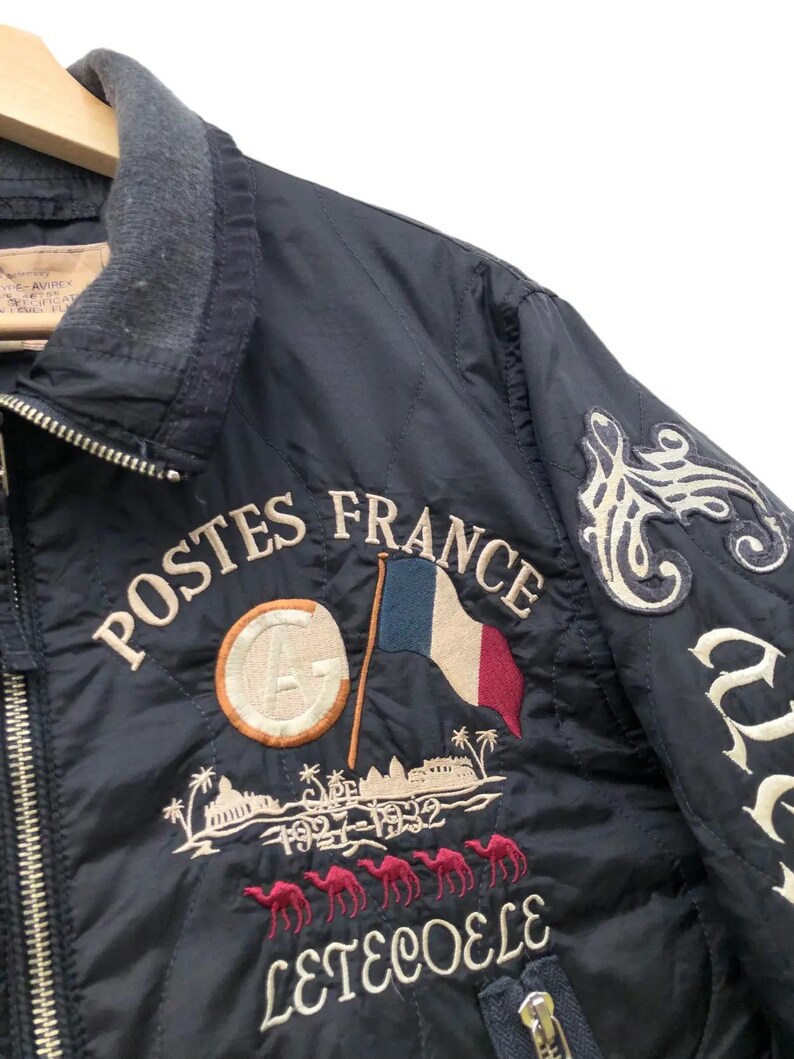 Puede incluir: Chaqueta acolchada azul marino con bordado "POSTES FRANCE", bandera francesa y figuras de camellos. Cuello gris y cierre de cremallera. Parches decorativos con letras blancas en la manga derecha.