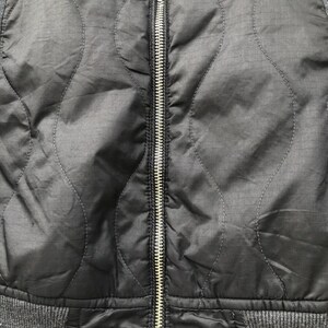 Puede incluir: Primer plano de una chaqueta negra acolchada con una cremallera plateada en el centro. La chaqueta tiene una parte inferior gris acanalada y tela gris en los hombros. El patr&oacute;n acolchado es una serie de l&iacute;neas curvas.