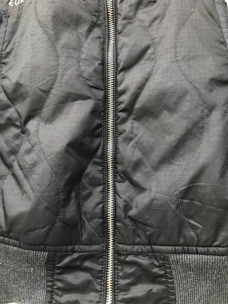 Puede incluir: Primer plano de una chaqueta negra acolchada con una cremallera plateada en el centro. La chaqueta tiene una parte inferior gris acanalada y tela gris en los hombros. El patr&oacute;n acolchado es una serie de l&iacute;neas curvas.